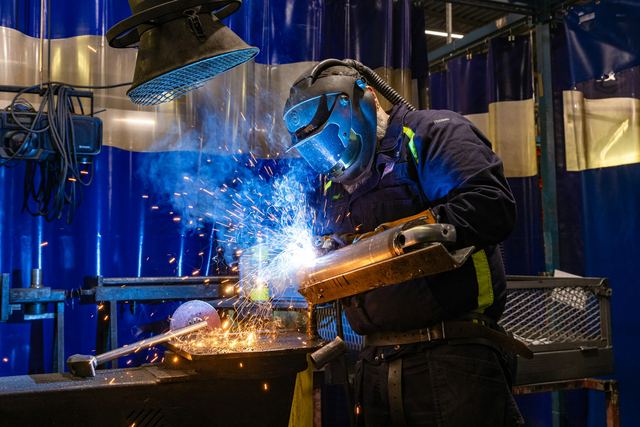 Soldador de máscara e luvas de proteção trabalhando numa peça de metal, cercado de faíscas, numa oficina