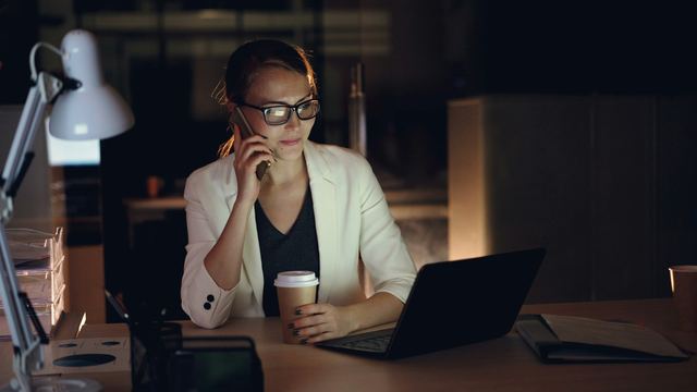 Mulher de óculos e blazer claro fala ao telefone sentada à mesa com notebook e copo de café, em um escritório escuro à noite.