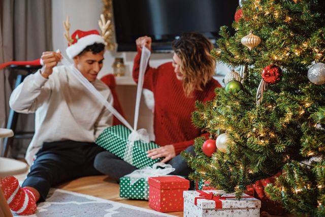 Casal sentado no chão abrindo presentes ao lado de uma árvore de Natal enfeitada; ele usa gorro de Papai Noel.