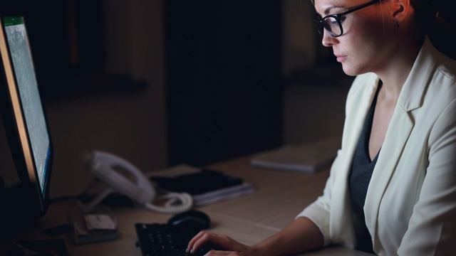 Mulher de óculos e blazer claro trabalhando no computador num escritório escuro, tarde da noite
