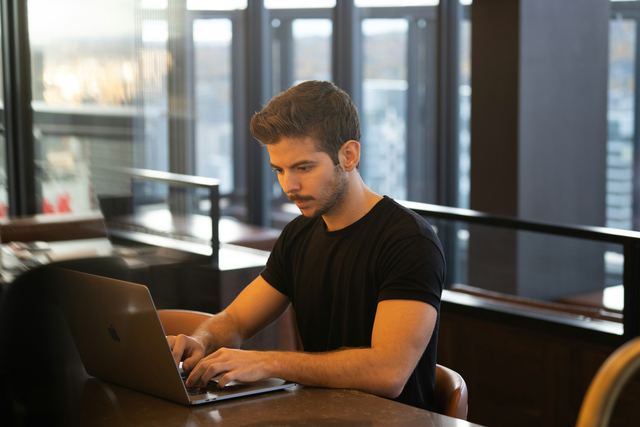 Homem de camiseta preta sentado em uma mesa digita em um notebook.