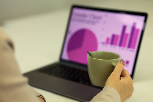 Pessoa de costas, com a mão numa caneca verde, olhando um notebook que mostra gráficos de pizza e de barras em tons de roxo