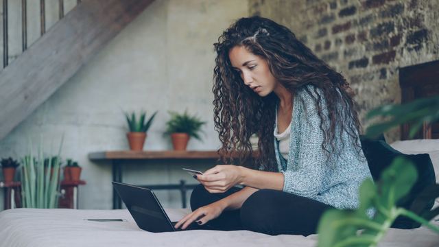 Mulher de cabelo cacheado sentada no chão da sala com um notebook e um cartão de crédito na mão, fazendo uma compra online.