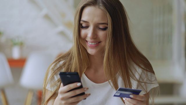 Jovem sorrindo em casa com o celular numa mão e o cartão na outra, comprando pela internet