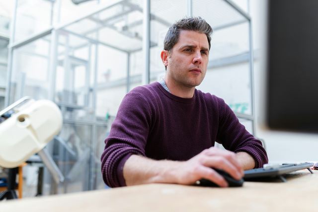 Homem de suéter roxo sentado à mesa trabalhando no computador