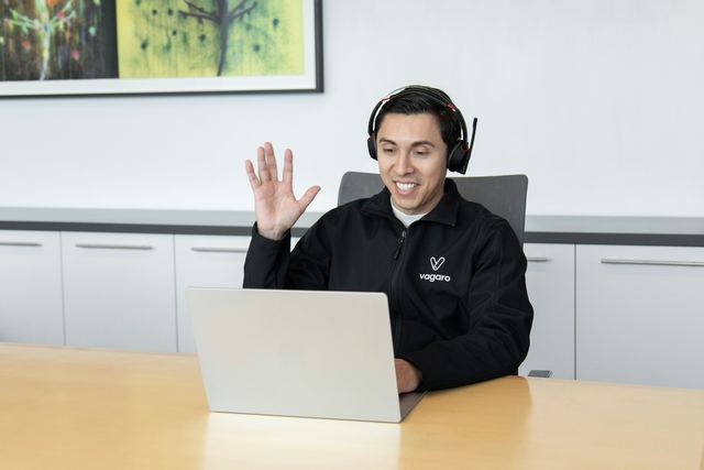 Atendente de fone de ouvido acenando sorridente para um cliente numa chamada pelo notebook