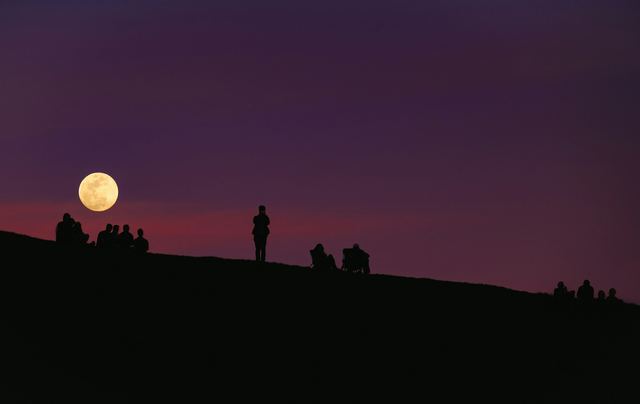 Grupo de pessoas em silhueta sob um céu roxo de noite com a lua ao fundo