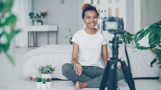 Jovem sentada no chão do quarto, sorrindo e falando para a câmera enquanto grava um vídeo