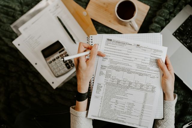 Pessoa conferindo documentos com uma calculadora e um notebook sobre a mesa