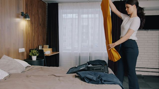 Mulher de blusa listrada dobrando um vestido amarelo para guardar na mala aberta sobre a cama, num quarto claro