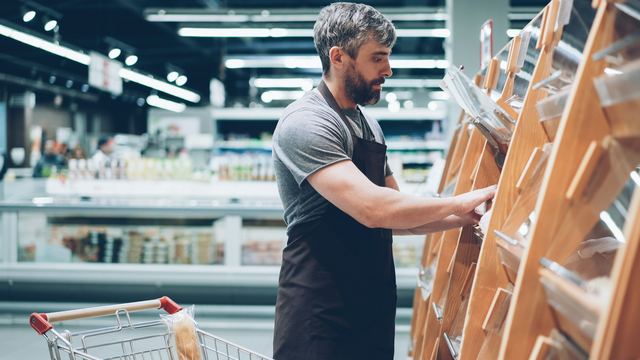 Funcionário de avental e barba repondo pães nas prateleiras da padaria de um mercado