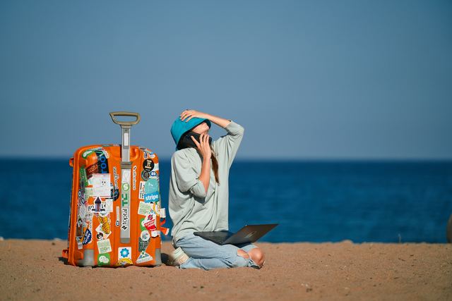 Mulher sentada na praia com uma mala e um notebook, trabalhando com o mar ao fundo