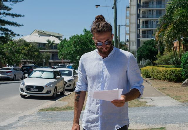Homem de camiseta branca segurando folhas de papel impresso, conferindo o boleto do imposto