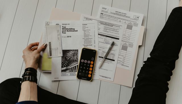 Pessoa segurando uma folha de papel perto de uma caneta e de uma calculadora, conferindo as contas na mesa