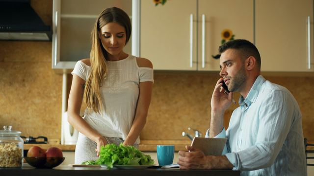 Mulher picando alface na cozinha enquanto um homem fala ao telefone atrás dela.