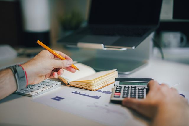 Pessoa sentada à mesa com uma calculadora e um caderno, fazendo as contas do mês