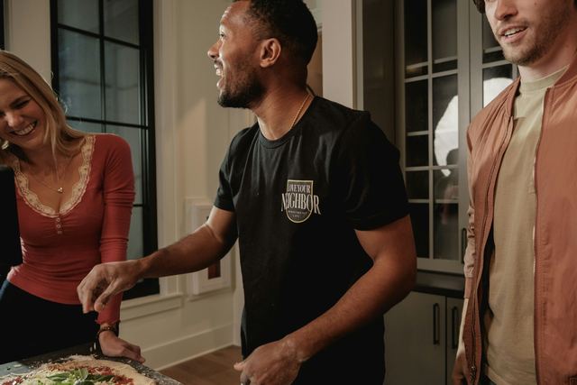 Amigos rindo na cozinha enquanto um deles coloca a cobertura na pizza