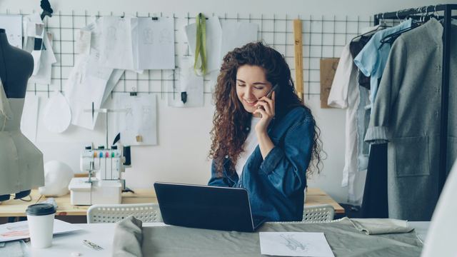 Dona de um ateliê de costura ao celular e ao notebook, com roupas e máquina de costura ao fundo