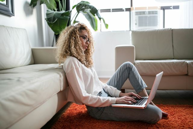Mulher sentada no chão, encostada no sofá, usando um notebook