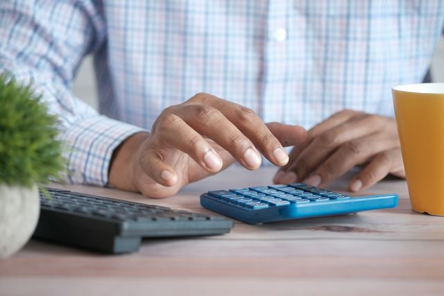 Mão de um homem usando uma calculadora sobre a mesa, ao lado de uma caneca de café