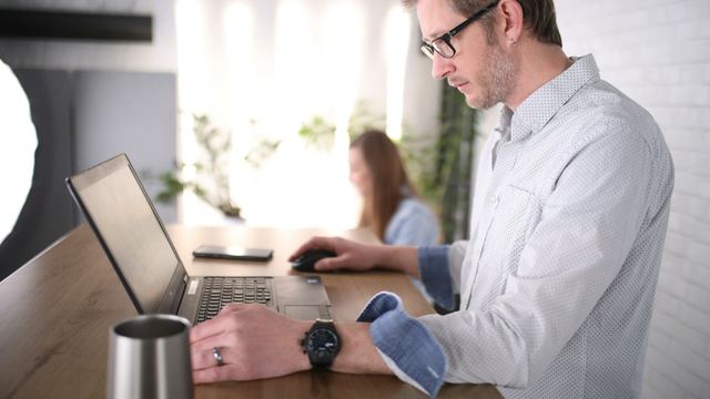 Homem de camisa branca trabalhando em pé diante de um computador em uma mesa de escritório