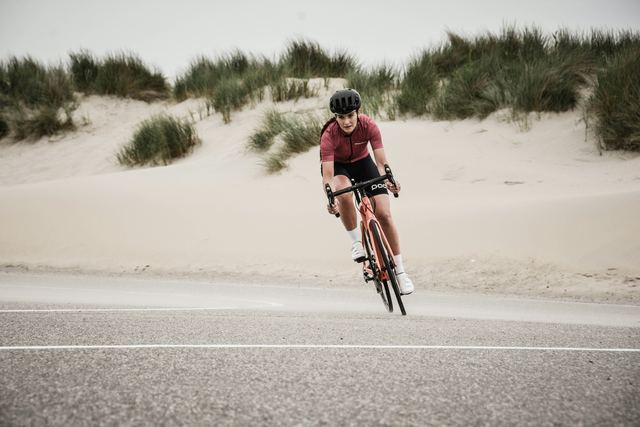 Ciclista de capacete e camisa vinho pedalando numa estrada à beira das dunas