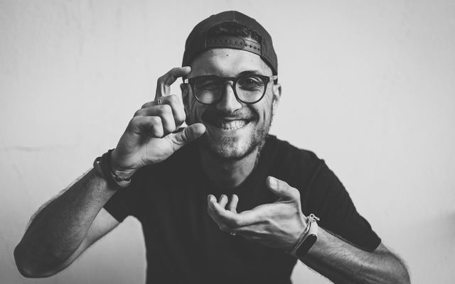 Homem de óculos e boné sorrindo, em preto e branco, com os dedos em pinça de uma mão como quem segura algo pequeno e a outra mão aberta.