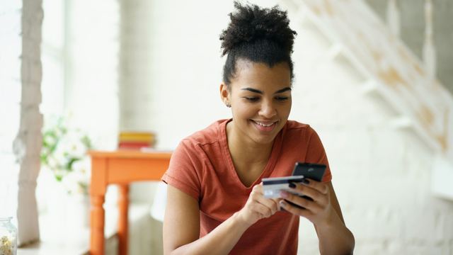 Jovem de cabelo cacheado sorrindo para o celular, com o cartão do banco na mão, numa casa clara