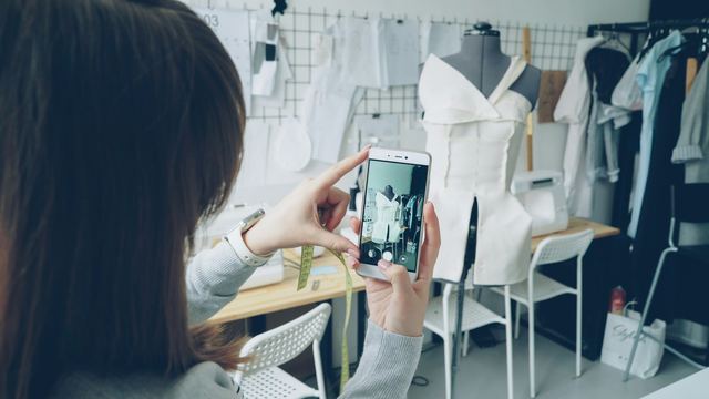Mãos fotografando com o celular uma peça de roupa montada num manequim, num ateliê de costura