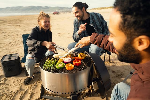 Três amigos rindo em volta de uma grelha na praia, com legumes e milho assando enquanto um deles vira a comida com o pegador