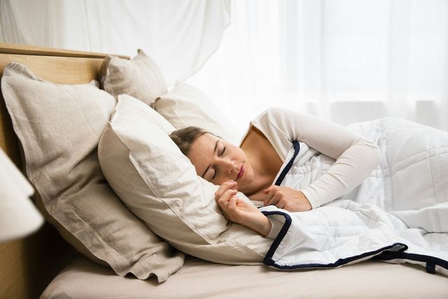 Mulher dormindo de lado em uma cama com edredom branco e travesseiros, com luz suave da janela.