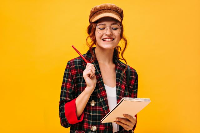Jovem sorridente de óculos, boina e jaqueta xadrez segurando um lápis e um caderno de espiral sobre fundo amarelo