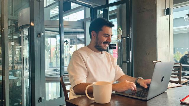 Homem sorrindo enquanto trabalha no notebook, num café de janelões, com uma caneca grande na mesa