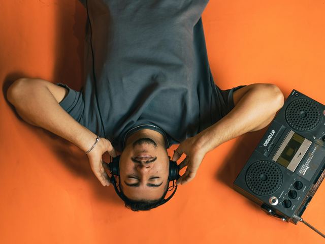 Homem deitado no chão laranja, fotografado de cima, de olhos fechados e fones de ouvido, ao lado de um rádio portátil antigo.