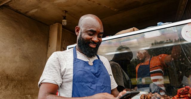 Homem de barba e avental azul, de camiseta clara, sorrindo para o celular à frente da vitrine de uma lanchonete