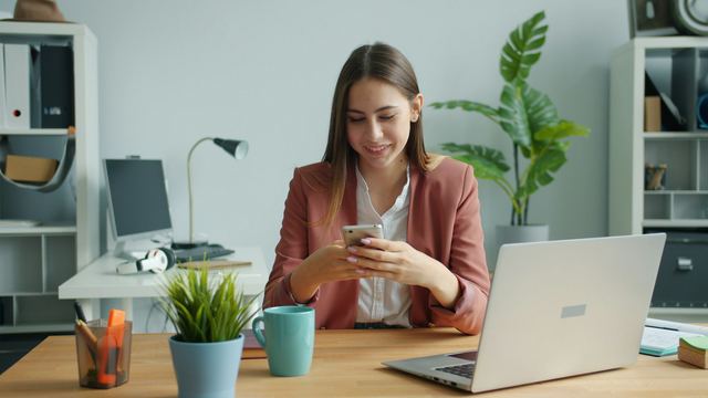 Jovem sorridente mexendo no celular à mesa de um escritório claro, com notebook, planta e caneca ao lado