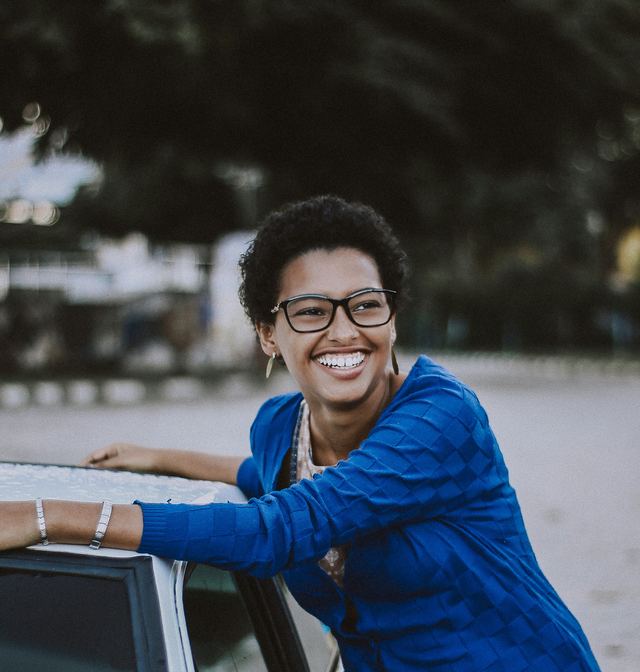Mulher de óculos e cardigã azul sorrindo, apoiada em um carro branco ao ar livre durante o dia