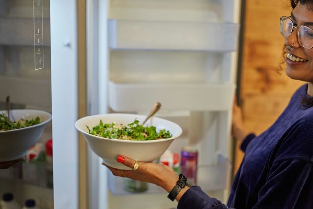 Mulher de óculos sorrindo enquanto guarda uma tigela de salada na geladeira aberta