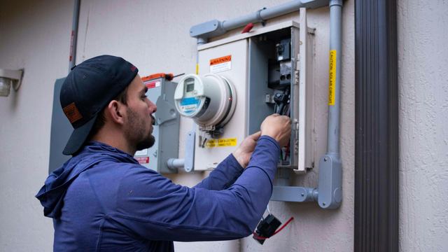 Homem de boné e moletom azul mexendo em um medidor de energia elétrica instalado na parede externa de uma casa.