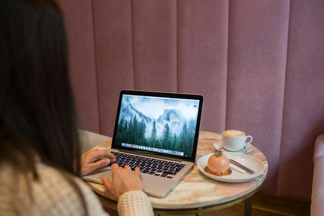 Mulher escrevendo no notebook numa mesa de café, com xícara e doce ao lado