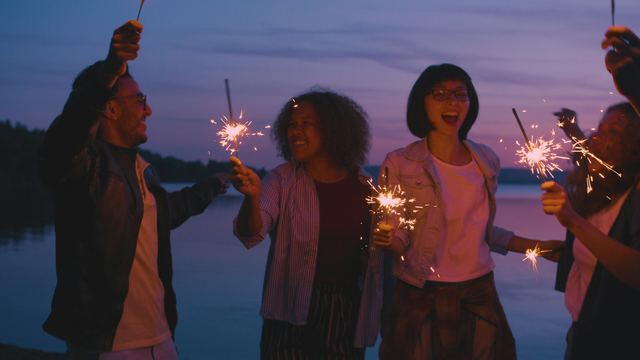 Grupo de amigos rindo e dançando ao entardecer à beira da água, segurando estrelinhas acesas.