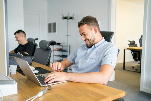 Homem de camisa azul-clara sorrindo enquanto digita em um notebook sobre uma mesa de madeira em um escritório claro