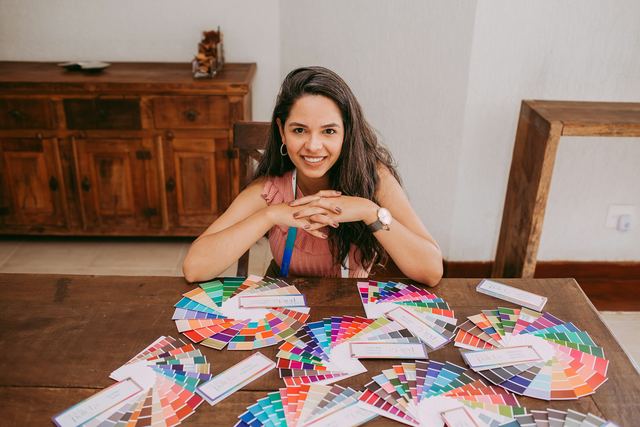Mulher sorridente sentada à mesa de madeira com os braços cruzados, cercada por leques de cores abertos.