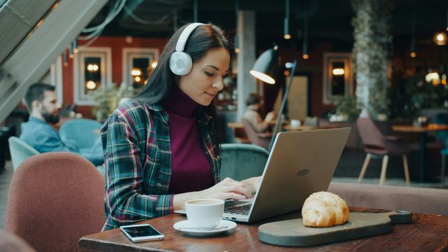 Mulher de fones de ouvido digitando no notebook numa mesa de café, com xícara e croissant ao lado