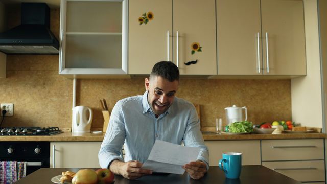 Homem sentado à mesa da cozinha lendo uma carta com atenção, durante o café da manhã