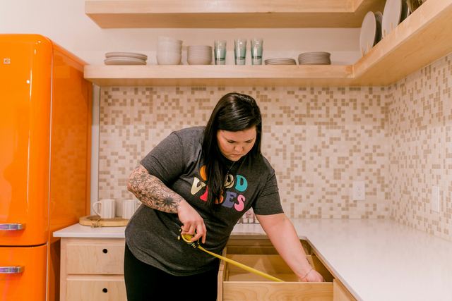 Mulher de camiseta cinza e tatuagem no braço medindo uma gaveta de cozinha com uma fita métrica.