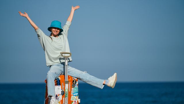 Viajante de chapéu azul sentada numa mala adesivada, de braços abertos, com o mar ao fundo