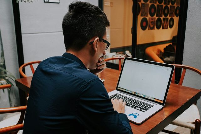 Homem de camisa escura sentado a uma mesa de madeira, de lado, digitando em um notebook.