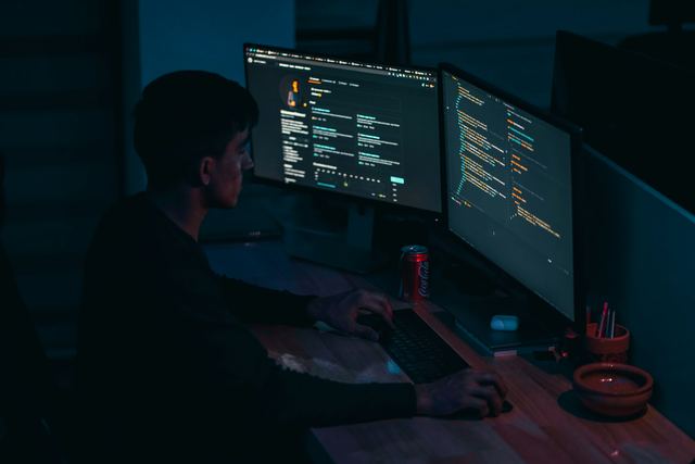 Desenvolvedor trabalhando à noite diante de dois monitores com código, num quarto escuro