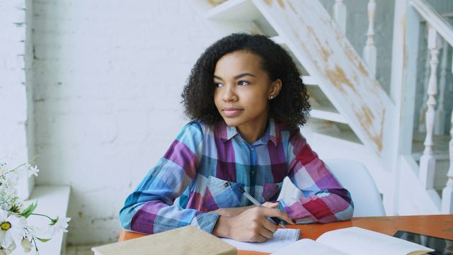 Estudante de cabelo cacheado escrevendo no caderno com um livro aberto na mesa